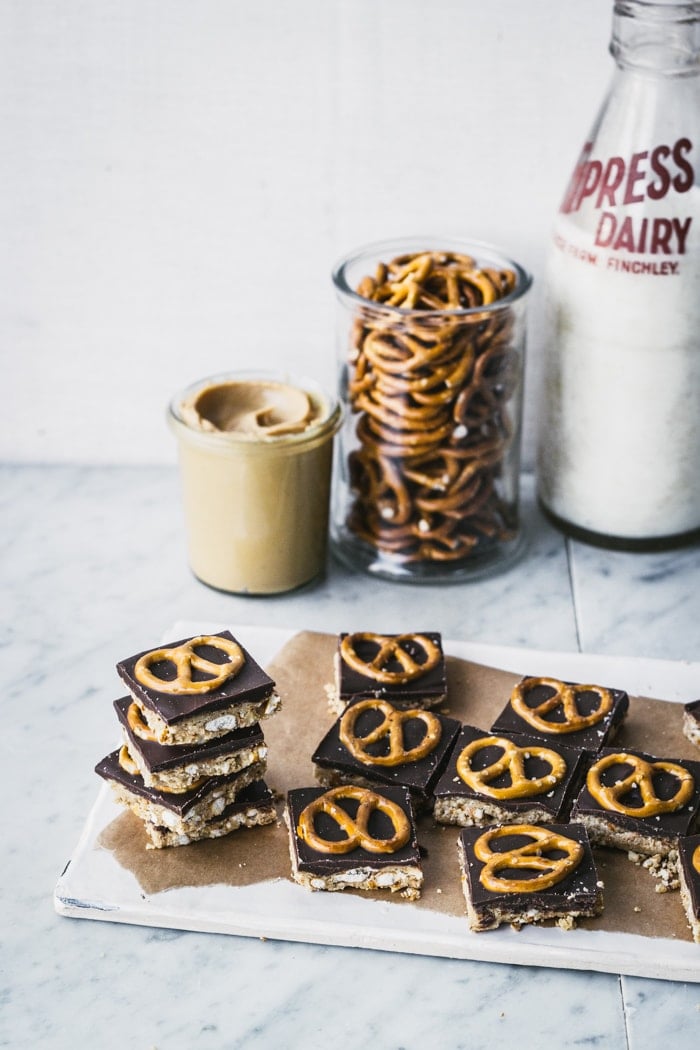 Chocolate peanut butter & pretzel bars stacked in a tower with jars of peanut butter, pretzels and milk behind